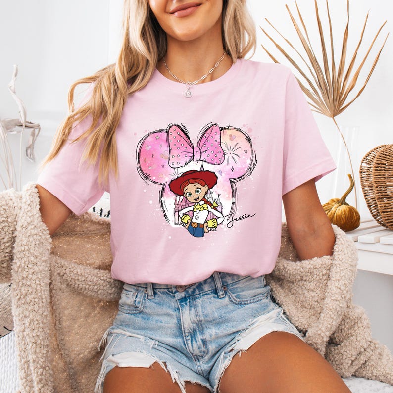 Puede incluir: Camiseta rosa con una imagen de dibujos animados de Jessie de Toy Story con un sombrero de vaquero y sosteniendo un lazo. La imagen est&aacute; dentro de una silueta de oreja de Mickey Mouse rosa con un lazo rosa. El texto "Jessie" est&aacute; escrito debajo de la imagen.