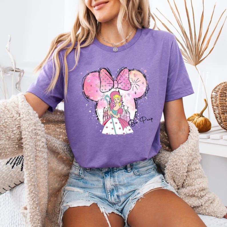 Puede incluir: Camiseta morada con un dise&ntilde;o de orejas de Mickey Mouse rosa y blanco con una imagen de dibujos animados de Bo Peep de Toy Story. El texto "Be Peep" est&aacute; escrito debajo de la imagen.