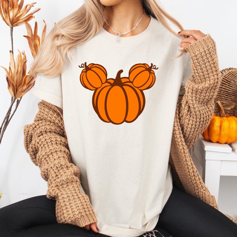 K&ouml;nnte beinhalten: Cremefarbenes T-Shirt mit einem orangefarbenen K&uuml;rbisdesign in Form eines M&auml;usekopfes. Das Design hat braune Umrisse. Das Shirt ist Teil einer Herbst- oder Halloween-Themenkollektion.