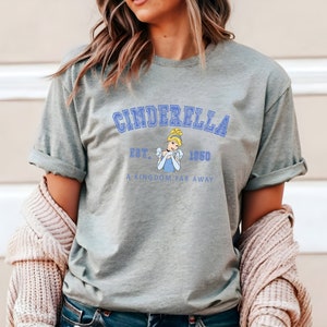 May include: A heather grey t-shirt with a blue graphic of Cinderella from the Disney movie Cinderella. The graphic includes the text "Cinderella Est. 1950 A Kingdom Far Away".