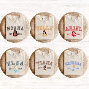 May include: Six white t-shirts with different Disney princess characters and names printed on them. Each shirt has a different princess, including Moana, Belle, Ariel, Elsa, Tiana, and Cinderella. The shirts also have the princess's name and the year they were established.