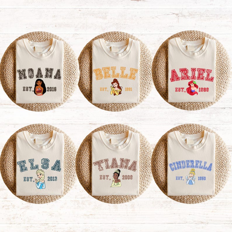 May include: Six white t-shirts with different Disney princess characters and names printed on them. Each shirt has a different princess, including Moana, Belle, Ariel, Elsa, Tiana, and Cinderella. The shirts also have the princess's name and the year they were established.