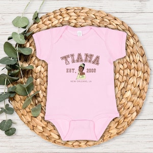 May include: Pink baby onesie with a brown graphic of Tiana from Disney's Princess and the Frog. The graphic includes the text "Tiana Est. 2003 New Orleans, LA".
