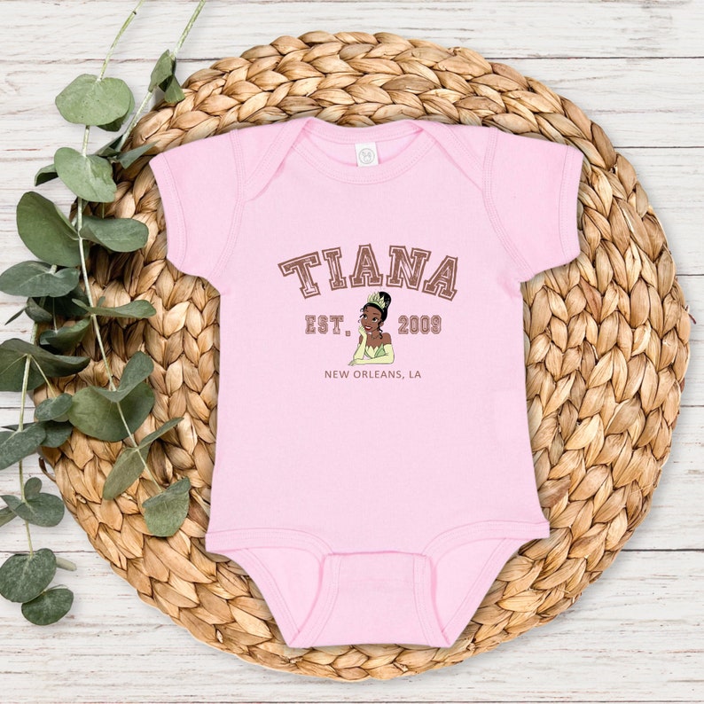 May include: Pink baby onesie with a brown graphic of Tiana from Disney's Princess and the Frog. The graphic includes the text "Tiana Est. 2003 New Orleans, LA".