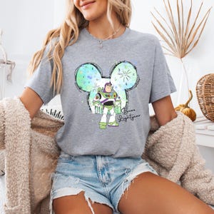 Puede incluir: Una camiseta gris con un dise&ntilde;o de cabeza de Mickey Mouse en acuarela verde, azul y blanco. El dise&ntilde;o presenta a Buzz Lightyear de Toy Story de pie dentro de la cabeza de Mickey Mouse. El texto "Buzz Lightyear" est&aacute; escrito debajo del dise&ntilde;o.