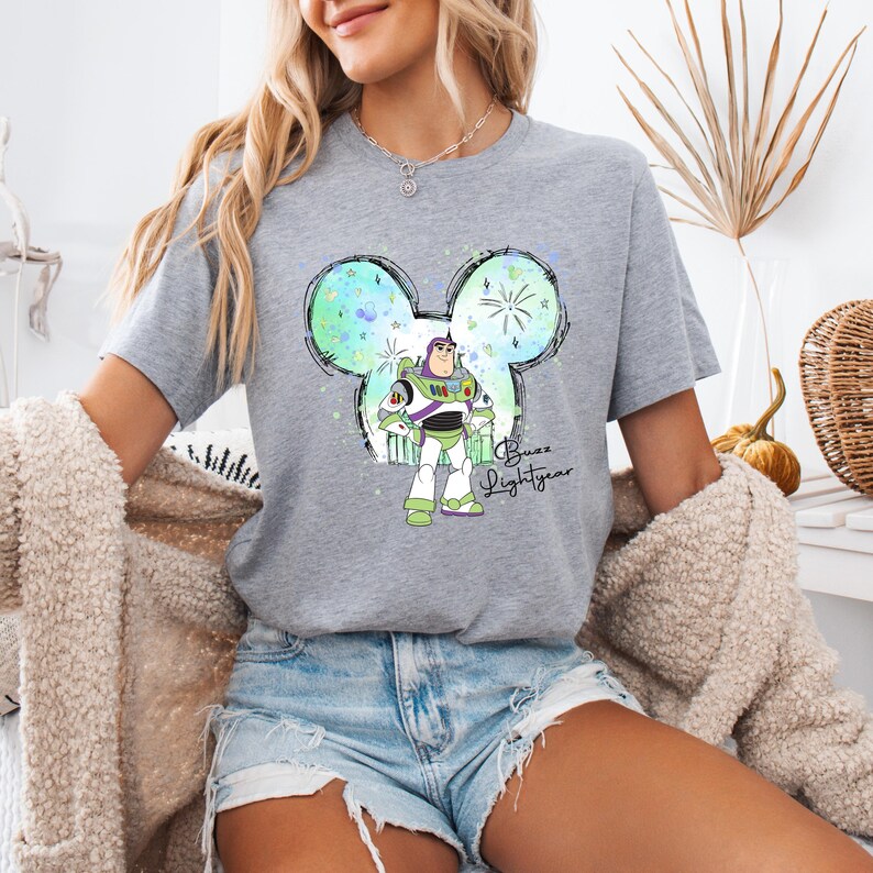Puede incluir: Una camiseta gris con un dise&ntilde;o de cabeza de Mickey Mouse en acuarela verde, azul y blanco. El dise&ntilde;o presenta a Buzz Lightyear de Toy Story de pie dentro de la cabeza de Mickey Mouse. El texto "Buzz Lightyear" est&aacute; escrito debajo del dise&ntilde;o.