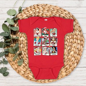 May include: Red baby onesie with a Christmas-themed design featuring nine Disney princesses in a grid pattern. Each princess is wearing a festive outfit and has her name written below her image. The princesses featured are Snow White, Ariel, Aurora, Cinderella, Tiana, Belle, Rapunzel, Elsa, and Jasmine.