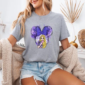 Pu&ograve; includere: Una maglietta grigia con un designo ad acquerello viola e bianco di una principessa Disney con un fiocco viola. La principessa ha lunghi capelli biondi e indossa un vestito viola.