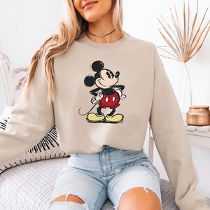 Puede incluir: Una sudadera beige con una imagen de dibujos animados de Mickey Mouse con un atuendo rojo y negro. La sudadera tiene mangas largas y cuello redondo.