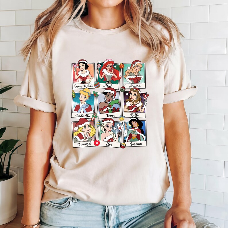 May include: A beige t-shirt with a graphic print of nine Disney princesses dressed in Christmas outfits. Each princess is in a separate frame with her name underneath. The princesses are Snow White, Ariel, Aurora, Cinderella, Tiana, Belle, Rapunzel, Elsa, and Jasmine.