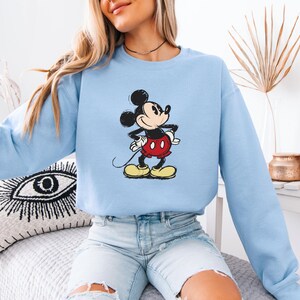 Puede incluir: Una sudadera de color azul claro con una imagen de dibujos animados de Mickey Mouse con un atuendo rojo y negro. Mickey Mouse est&aacute; de pie con la mano derecha en la cadera y la mano izquierda sosteniendo su cola.