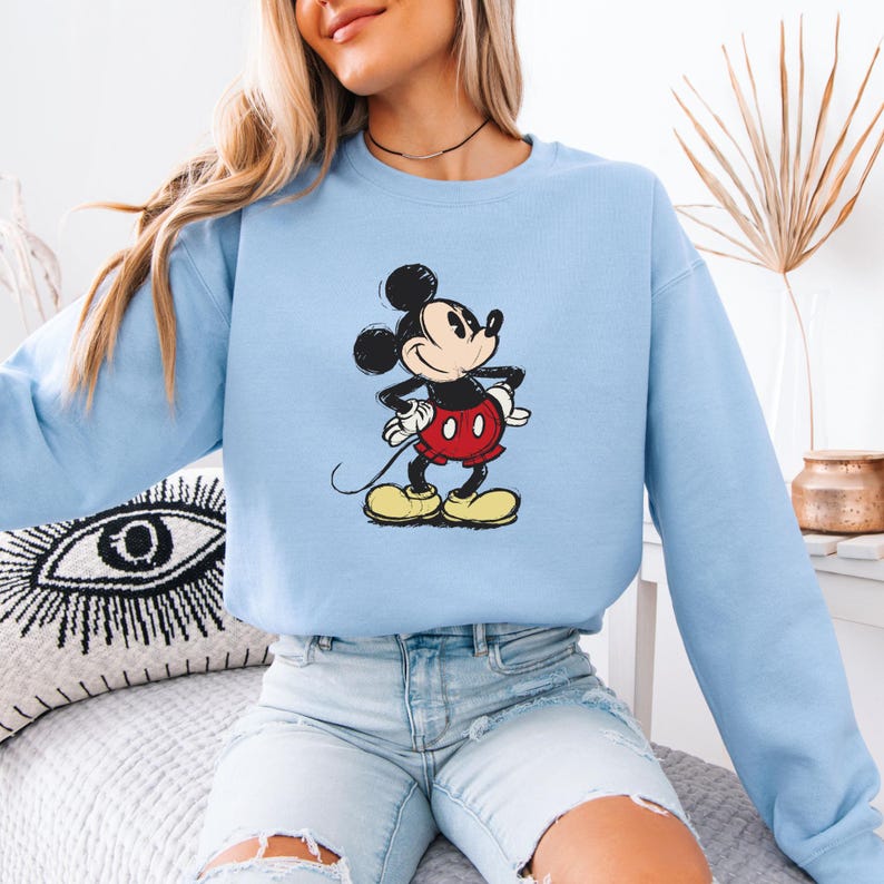 Puede incluir: Una sudadera de color azul claro con una imagen de dibujos animados de Mickey Mouse con un atuendo rojo y negro. Mickey Mouse est&aacute; de pie con la mano derecha en la cadera y la mano izquierda sosteniendo su cola.