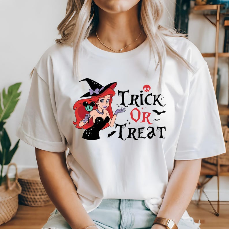 May include: White t-shirt with a graphic of a cartoon mermaid wearing a witch hat and holding a Mickey Mouse lollipop. The text "Trick or Treat" is printed below the image.