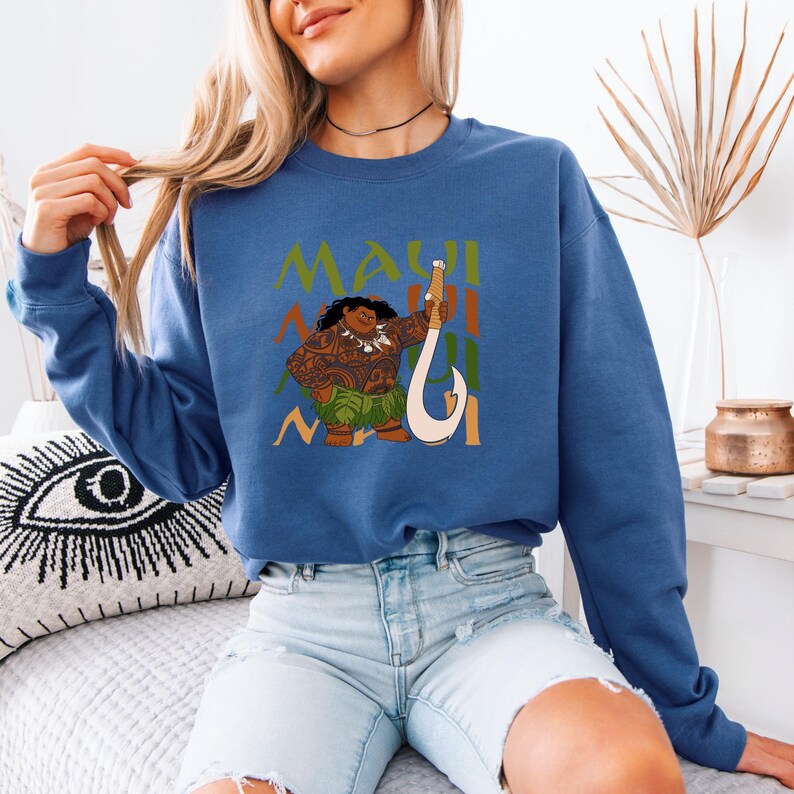 Puede incluir: Una sudadera azul con un gr&aacute;fico de Maui de la pel&iacute;cula Moana. El gr&aacute;fico presenta a Maui sosteniendo su anzuelo de pesca y la palabra "Maui" se repite en una fuente estilizada.