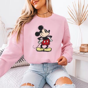 Puede incluir: Una sudadera rosa con una imagen de dibujos animados de Mickey Mouse en blanco y negro. Mickey Mouse lleva pantalones cortos rojos y zapatos amarillos.