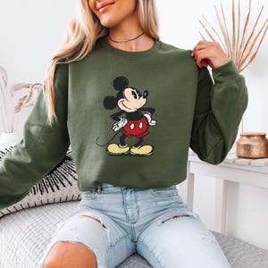 Puede incluir: Una sudadera verde con una imagen de dibujos animados de Mickey Mouse en negro y rojo. Mickey Mouse est&aacute; sonriendo y tiene las manos en las caderas.