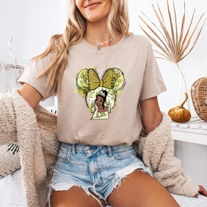 Pu&ograve; includere: Una maglietta beige chiaro con una grafica di una principessa dei cartoni animati con una corona e un vestito verde. La principessa indossa un fiocco d'oro ed &egrave; circondata da un design acquerellato giallo e verde. Il design ha la forma delle orecchie di Topolino.