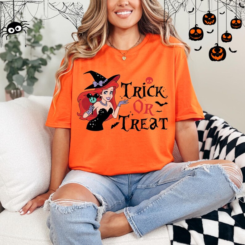 May include: Orange t-shirt with a graphic of a witch wearing a black dress and a witch hat. The witch is holding a lollipop and has red hair. The text "Trick or Treat" is printed on the shirt.