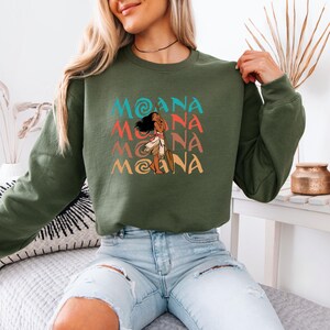 Puede incluir: Una sudadera verde con un estampado gr&aacute;fico de Moana de la pel&iacute;cula de Disney Moana. El gr&aacute;fico es un patr&oacute;n repetido de la palabra "Moana" en diferentes colores y una imagen de dibujos animados de Moana sosteniendo un remo.
