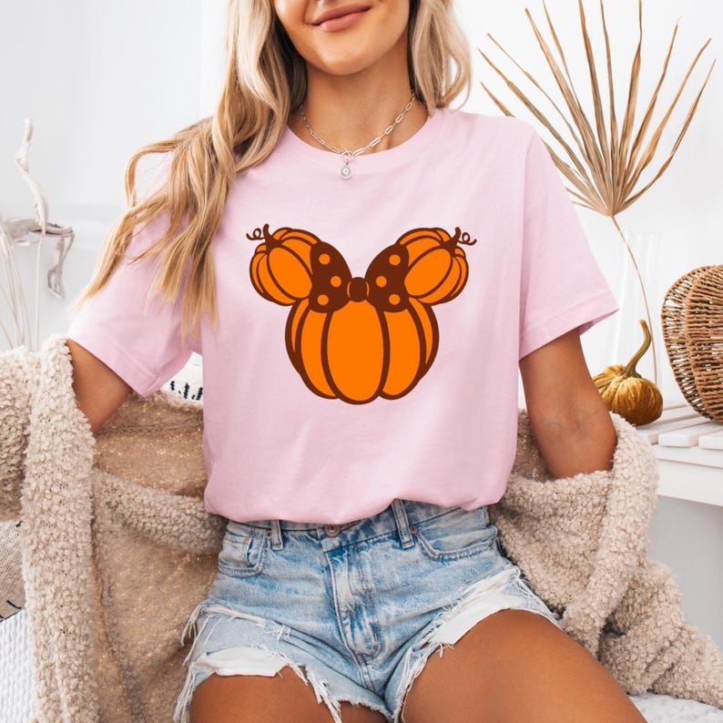 K&ouml;nnte beinhalten: Ein hellrosa T-Shirt mit einem K&uuml;rbis-Design. Das Design ist ein orangefarbener K&uuml;rbis mit einer braunen Schleife und einem Stiel, der an den Kopf einer Figur erinnert. Das Shirt hat einen Rundhalsausschnitt und kurze &Auml;rmel.