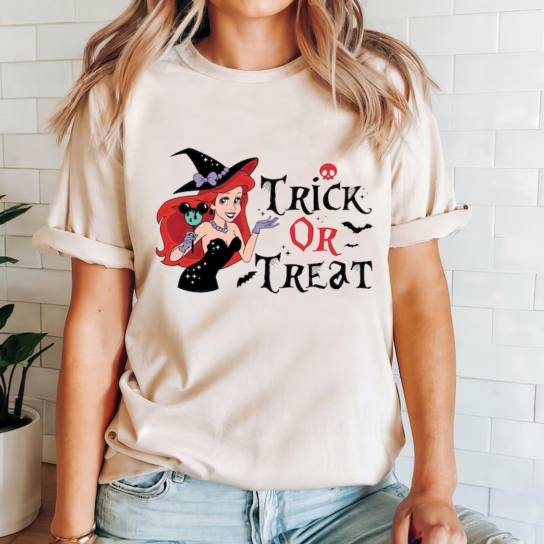 May include: A white t-shirt with a black and red graphic of a witch dressed as Ariel from The Little Mermaid. The graphic includes the text "Trick or Treat" with bats and a skull.