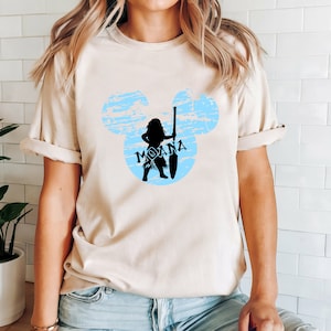 May include: A light beige t-shirt with a blue and white Mickey Mouse silhouette featuring a black silhouette of Moana holding a spear. The text "Moana" is written in black inside the silhouette.