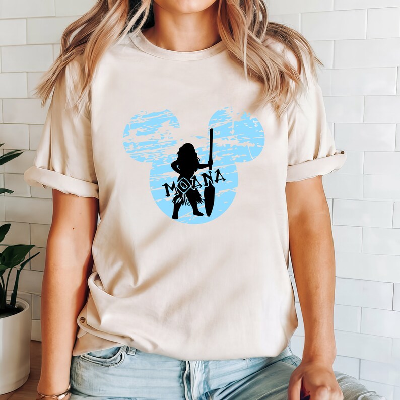 May include: A light beige t-shirt with a blue and white Mickey Mouse silhouette featuring a black silhouette of Moana holding a spear. The text "Moana" is written in black inside the silhouette.