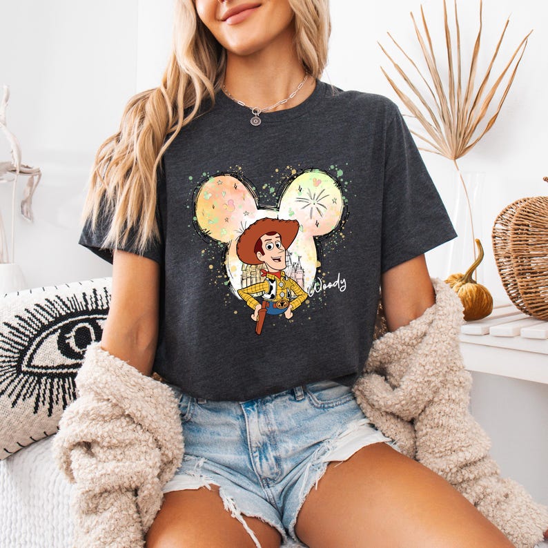 Puede incluir: Una camiseta gris oscuro con una silueta de Mickey Mouse de estilo acuarela. Dentro de la silueta hay una imagen de dibujos animados de Woody de Toy Story. El texto "Woody" est&aacute; escrito en blanco debajo de la imagen.