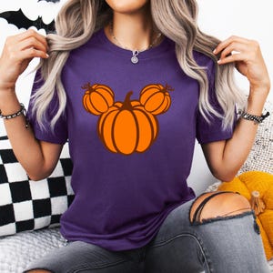 K&ouml;nnte beinhalten: Ein lila T-Shirt mit einem Halloween-Design. Die Grafik zeigt einen stilisierten orangefarbenen K&uuml;rbis mit zwei kleineren K&uuml;rbissen dar&uuml;ber, die an den Kopf einer ber&uuml;hmten Maus erinnern. Das T-Shirt besteht aus weichem Material und eignet sich f&uuml;r Freizeitkleidung.