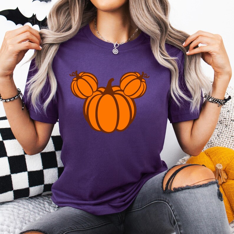 K&ouml;nnte beinhalten: Ein lila T-Shirt mit einem Halloween-Design. Die Grafik zeigt einen stilisierten orangefarbenen K&uuml;rbis mit zwei kleineren K&uuml;rbissen dar&uuml;ber, die an den Kopf einer ber&uuml;hmten Maus erinnern. Das T-Shirt besteht aus weichem Material und eignet sich f&uuml;r Freizeitkleidung.