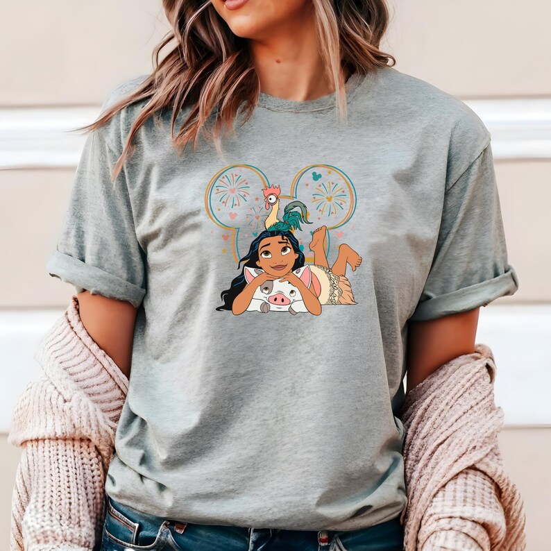 Może przedstawiać: Szary T-shirt z kresk&oacute;wkową postacią Moany z filmu Disneya Moana, z jej świnią Pua i kogutkiem Hei Hei. Obrazek znajduje się w sylwetce ucha Myszki Miki z fajerwerkami.