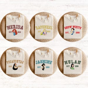 May include: Six white t-shirts with different Disney princess characters and names printed on them. The shirts have a crew neck and short sleeves. The names are Merida, Rapunzel, Snow White, Pocahontas, Jasmine, and Mulan. Each shirt has an established date printed below the name.