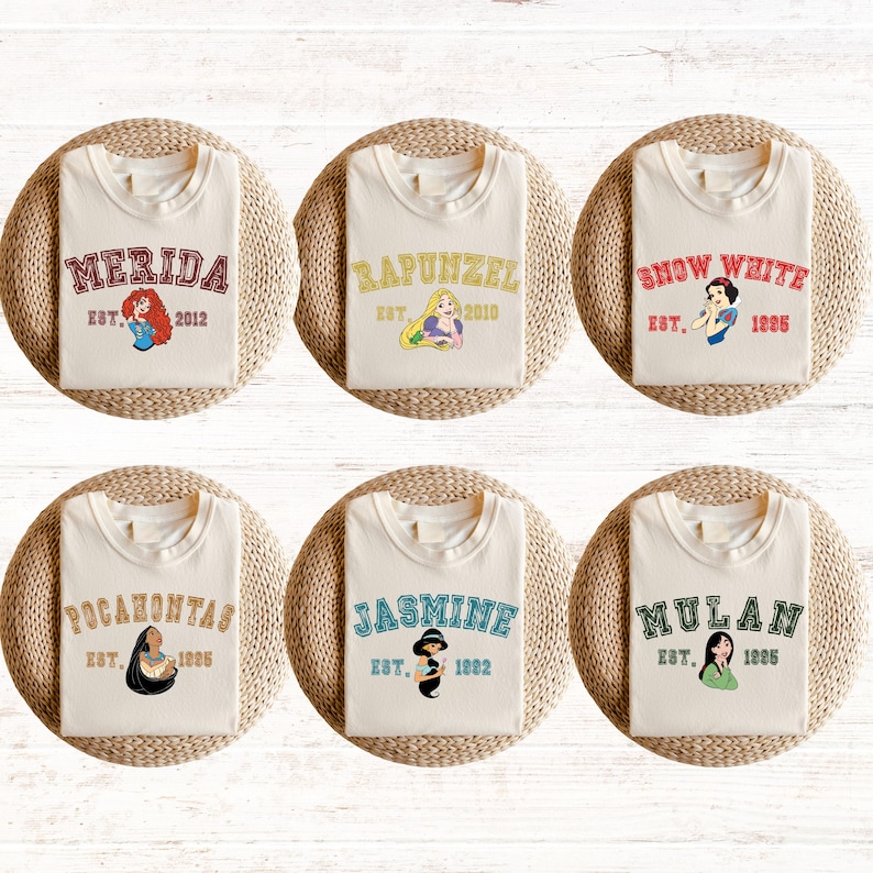 May include: Six white t-shirts with different Disney princess characters and names printed on them. The shirts have a crew neck and short sleeves. The names are Merida, Rapunzel, Snow White, Pocahontas, Jasmine, and Mulan. Each shirt has an established date printed below the name.