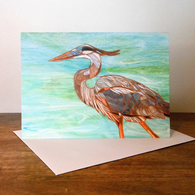 May include: A greeting card featuring a mosaic-style illustration of a blue heron standing in a turquoise and green water background.