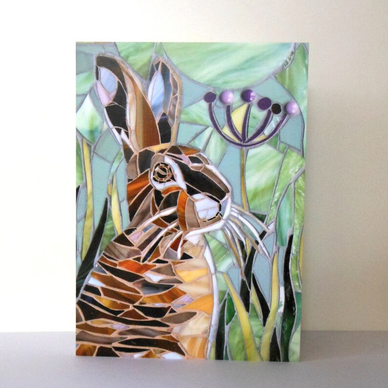 May include: A mosaic artwork depicting a brown and white rabbit in a green field with purple flowers. The rabbit is looking to the right and has long whiskers. The mosaic is made of small pieces of glass and tile.