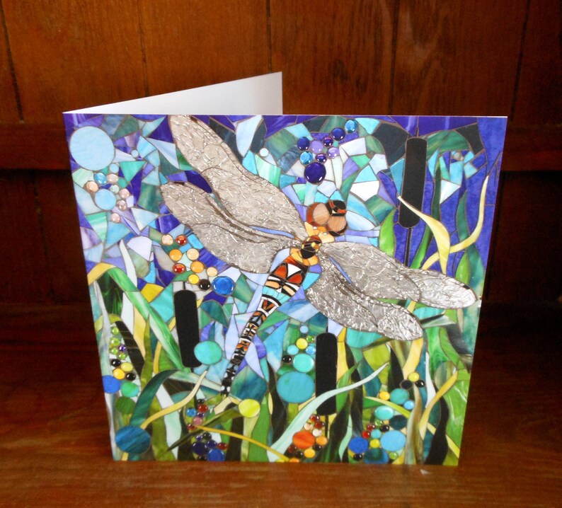 K&ouml;nnte beinhalten: Eine Gru&szlig;karte mit einer Libelle aus Mosaik mit silbernen Fl&uuml;geln, umgeben von blauen, gr&uuml;nen und gelben Mosaikdetails. Die Libelle ist vor einem Hintergrund aus blauem und violettem Mosaik platziert.