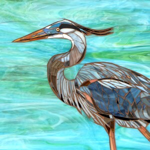 May include: A mosaic art piece depicting a blue heron standing in turquoise water. The heron is made of small pieces of glass and metal, creating a textured and detailed image.