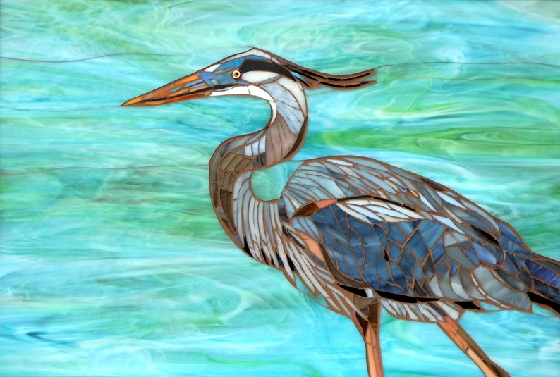 May include: A mosaic art piece depicting a blue heron standing in turquoise water. The heron is made of small pieces of glass and metal, creating a textured and detailed image.