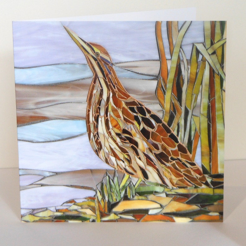 May include: A mosaic art piece depicting a brown and tan American bittern bird standing in reeds. The bird is made of small pieces of glass and tile, creating a textured and colorful image.