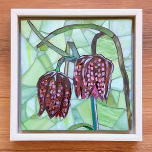 May include: A square mosaic art piece framed in light wood, depicting two drooping checkered fritillary flowers in shades of deep purple and brown against a background of various light green glass shards arranged in a geometric pattern.