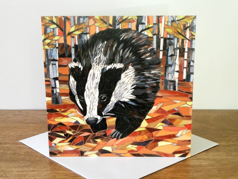 May include: A mosaic card featuring a badger in an autumnal forest setting. The badger is made of small, colorful tiles and is walking through a forest of birch trees. The background is a mix of orange, brown, and yellow tiles, representing fallen leaves.