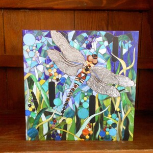 K&ouml;nnte beinhalten: Ein Mosaik-Kunstwerk, das eine Libelle mit silbernen Fl&uuml;geln vor einem blauen und gr&uuml;nen Hintergrund zeigt. Die Libelle ist von Schilf und anderen Wasserpflanzen umgeben.