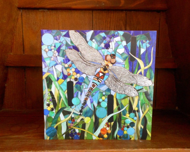 K&ouml;nnte beinhalten: Ein Mosaik-Kunstwerk, das eine Libelle mit silbernen Fl&uuml;geln vor einem blauen und gr&uuml;nen Hintergrund zeigt. Die Libelle ist von Schilf und anderen Wasserpflanzen umgeben.