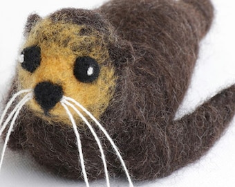 May include: A handmade felt sea otter figurine. The otter has a brown body, a tan face, and black eyes. White whiskers extend from the face. The otter is lying down with its arms at its sides. The background is white.