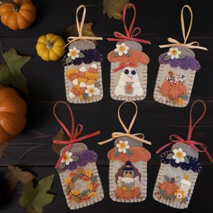 May include: Handmade felt jar ornaments with autumn themes. Each ornament features a unique design, including candy corn, a ghost, pumpkins, and a turkey. The ornaments have colorful accents and ribbon hangers, perfect for fall decor.