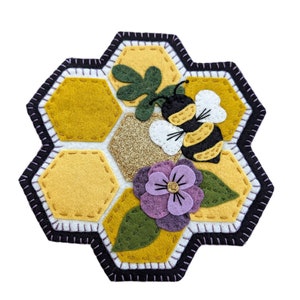 May include: A felt bee and flower appliqué with a honeycomb background. The bee is black and yellow with white wings. The flower is purple with a yellow centre. The honeycomb is yellow with a gold glitter centre.