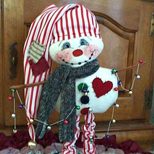 May include: A whimsical snowman doll with a striped red and white hat and legs. The snowman has a gray scarf, a red heart, and a string of lights. The snowman's black shoes have small gold bells. A festive holiday decoration.