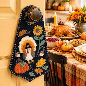 May include: A dark navy blue felt door hanger featuring a central applique of a turkey wearing a black hat, surrounded by embroidered sunflowers, orange pumpkins, and autumn leaves with visible white blanket stitching along the edges.