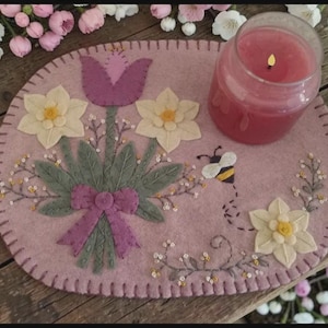 May include: A pink felt floral placemat with a bouquet of daffodils and a purple tulip. A lit pink candle in a glass jar sits on the mat. A felt bee flies near a daffodil.