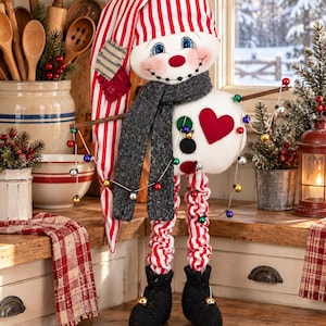 May include: A fabric snowman figurine wearing a red and white striped nightcap, a dark gray knit scarf, and black boots. The snowman features a red heart patch on its chest, twig arms holding a string of colorful miniature lights, and long legs.
